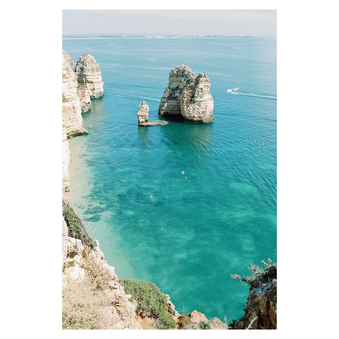Auto-generated description: Clear turquoise waters surround rugged rock formations near a coastline, with a boat and a few swimmers visible.