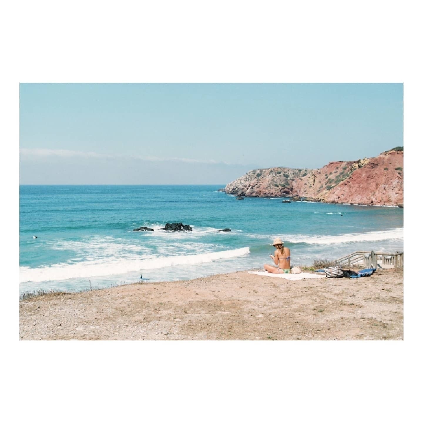 Auto-generated description: A person relaxes on a towel overlooking a beach with clear blue water and rocky cliffs.
