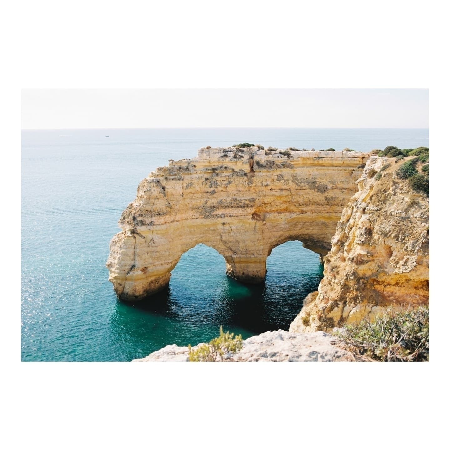 Auto-generated description: A natural rock formation with arches rises from the calm, blue ocean waters along a rocky coastline.