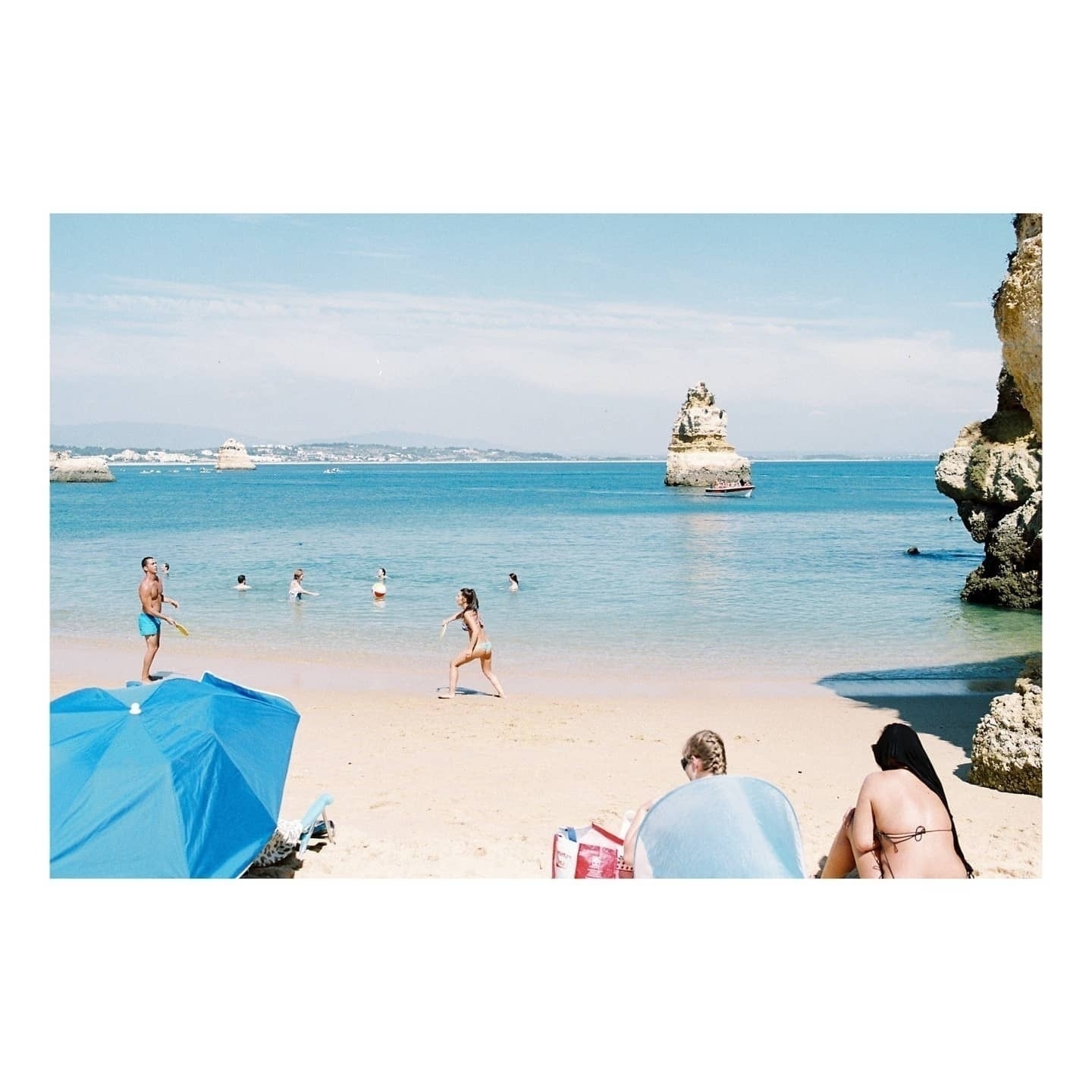 Auto-generated description: People are enjoying a sunny day at the beach, swimming and relaxing by the shore with blue umbrellas nearby.