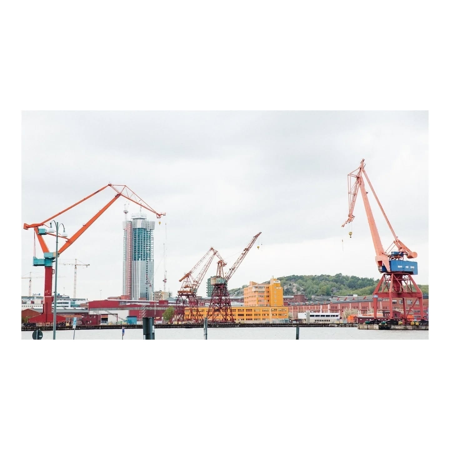 Auto-generated description: Cranes and industrial structures stand prominently against a cloudy sky, with a tall building and harbor in the background.