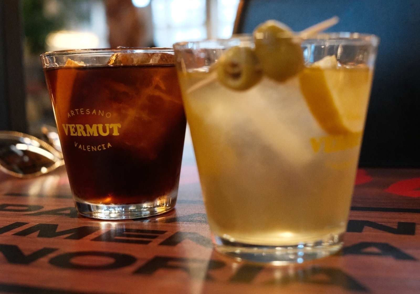 Auto-generated description: Two glasses filled with different kinds of vermouth, garnished with olives and a lemon slice, are placed on a table.